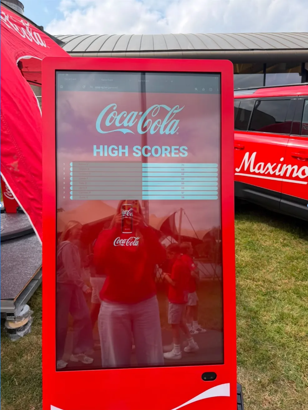 Het scorebord in de activatie: een verticaal scherm in een Coca-Cola-totem, hier op een fandag. Vol zonlicht, spiegelend glas, en toch leesbaar. Deelnemersnamen afgedekt.